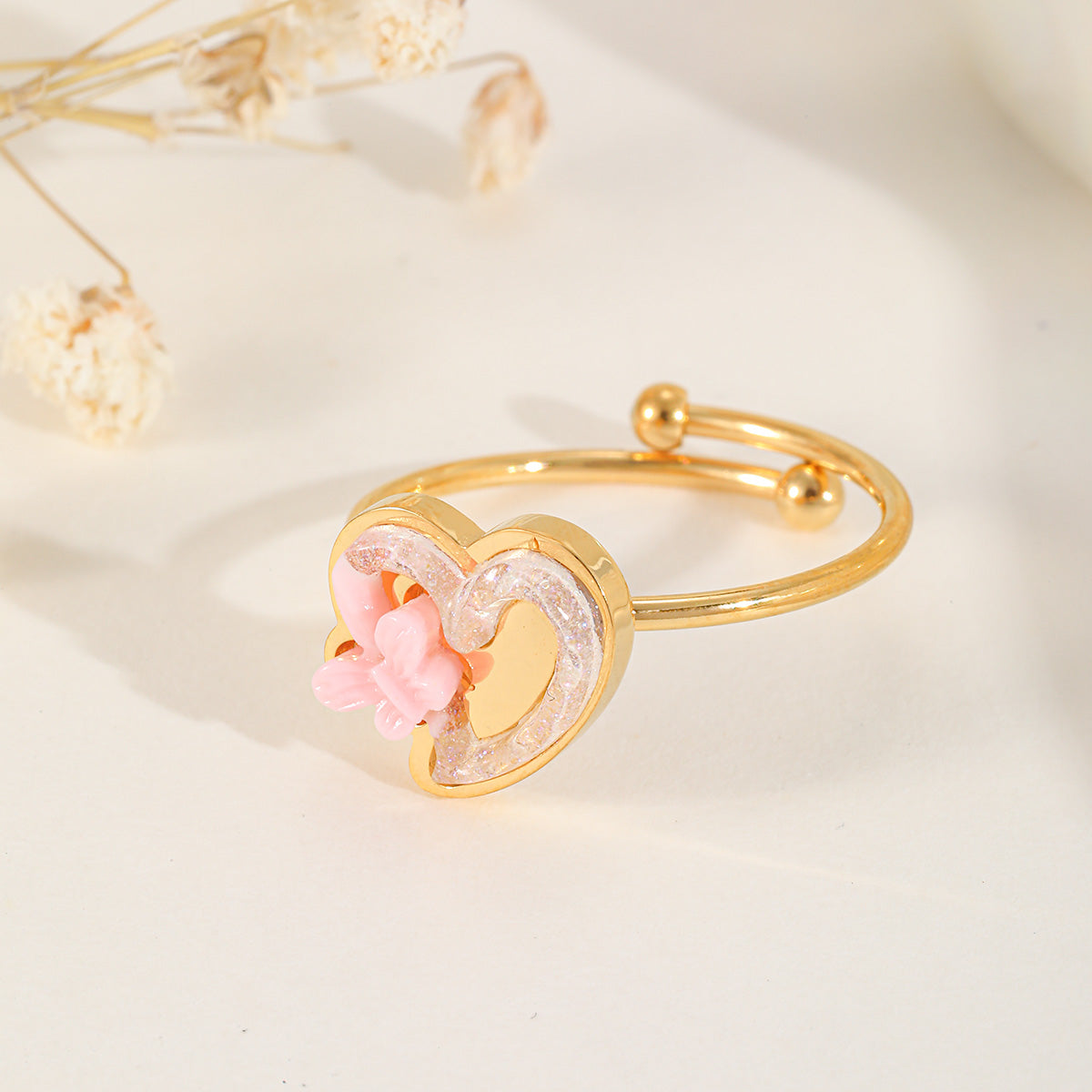 A gold adjustable ring featuring a pink heart charm with a smaller pink flower detail and shimmering resin border.