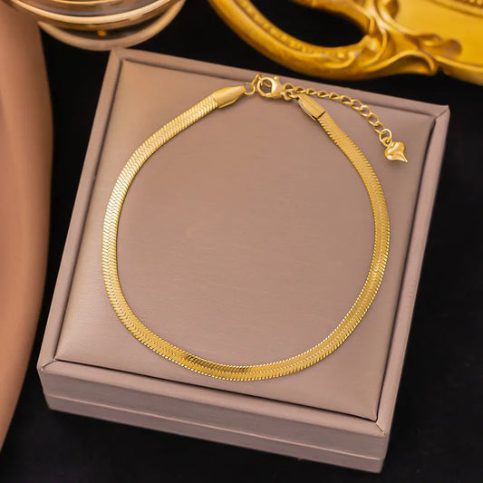 A 4mm wide gold herringbone flat snake chain bracelet with a heart-shaped charm on the extender, presented on a jewelry gift box.
