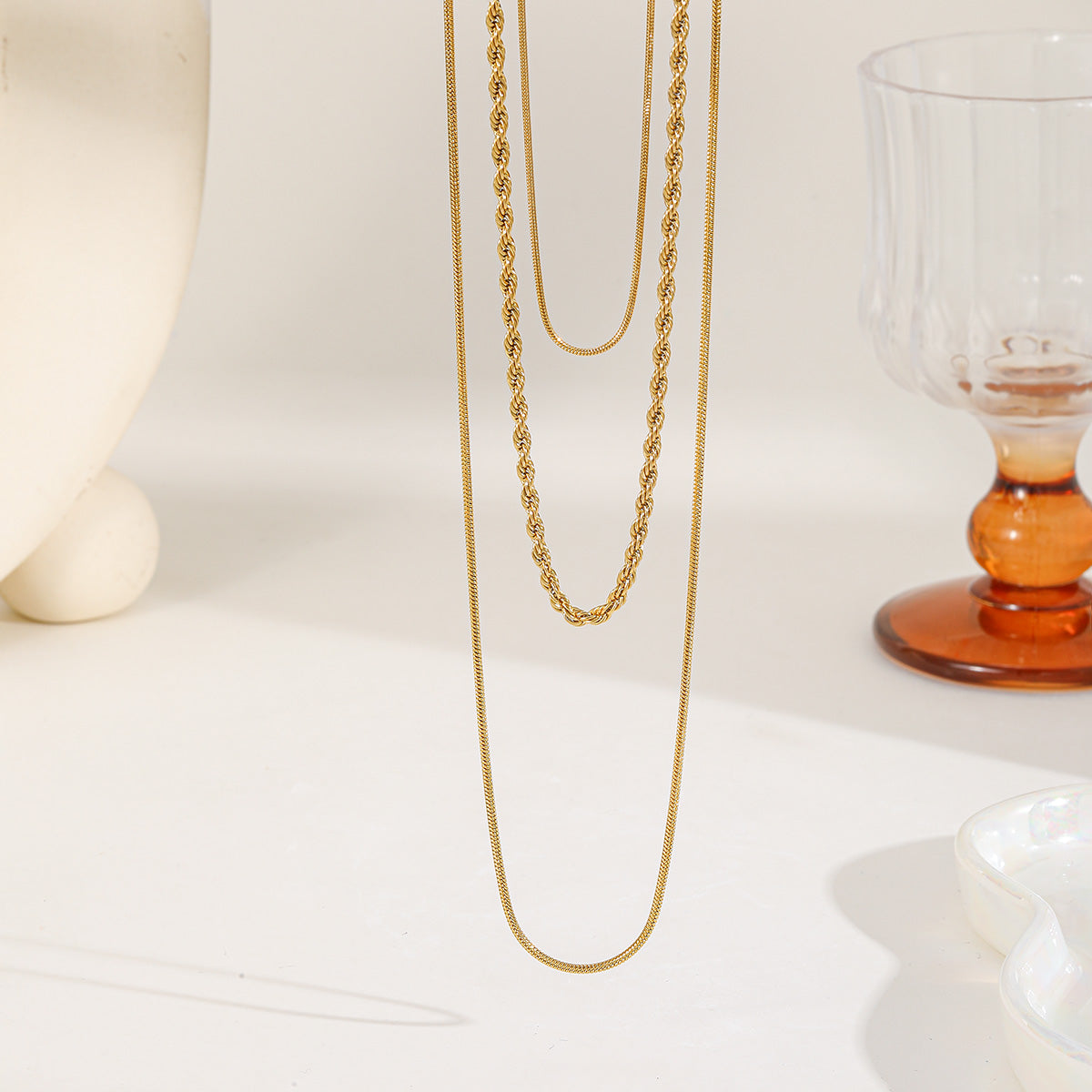 Close up of a three-layer gold necklace set featuring two thin snake chains and one twisted rope chain on a white minimalist background
