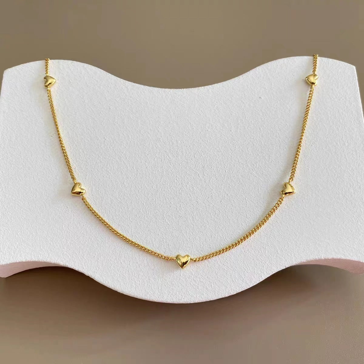 Full view of the dainty gold heart station necklace displayed on a white textured stand.