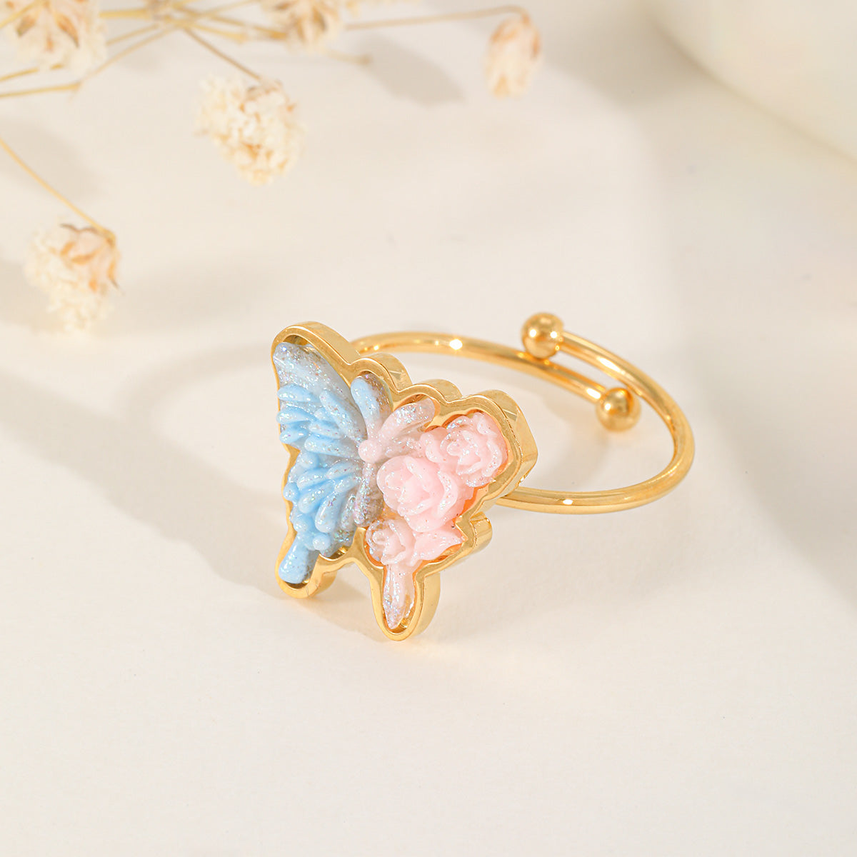 Close-up of a romantic gold ring with a large butterfly charm featuring intricate pink and blue shimmering resin details.