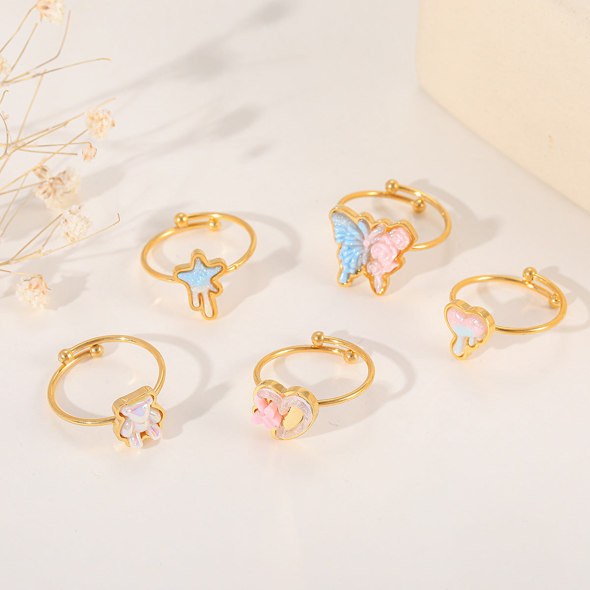 A collection of five gold adjustable rings with various resin charms, including a butterfly, heart, star, and bear, displayed on a white surface with dried flowers.