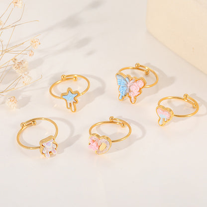 A collection of five gold adjustable rings with various resin charms, including a butterfly, heart, star, and bear, displayed on a white surface with dried flowers.