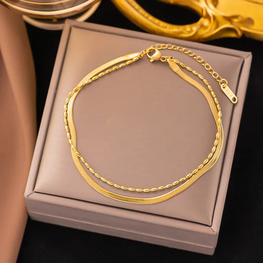 A double-layer gold-plated bracelet featuring a flat snake chain and a delicate bead chain, displayed on a taupe jewelry box.